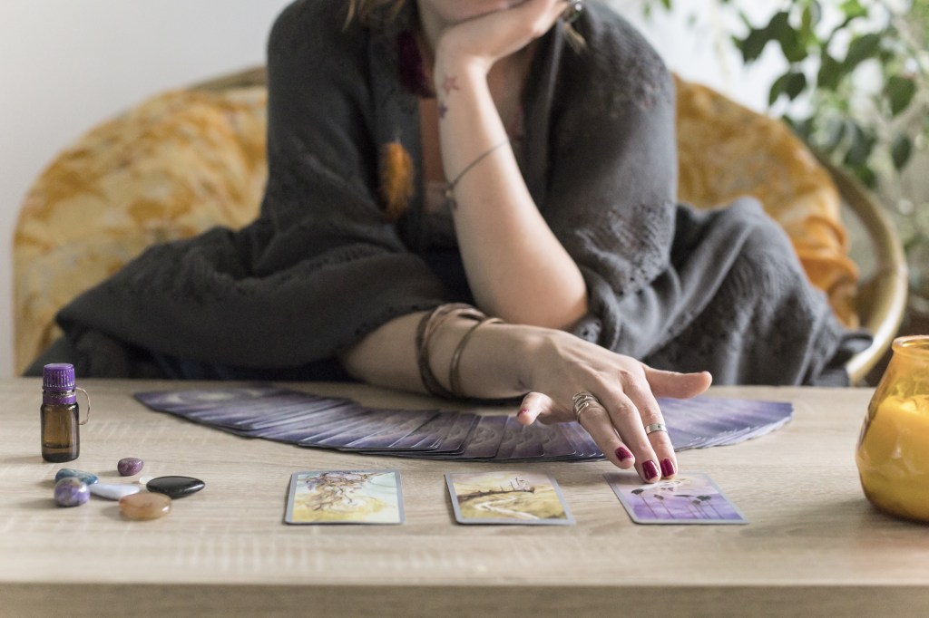 A white woman, visible from the chin down, sits behind a wooden table with tarot cards spread out in front of her. 