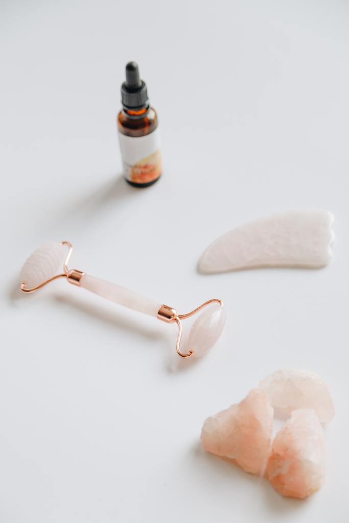 A bottle of essential oils, a rose quartz facial roller, and peach/pink crystals sit on a white surface.